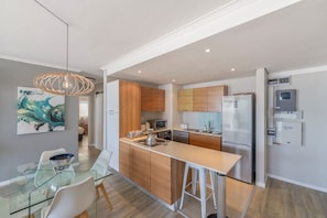 A modern kitchen with wooden cabinets, a stainless steel refrigerator, and a glass dining table with chairs. - Horizon Bay 903 Fresh Beachfront Hideaway (Bloubergstrand)