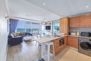 A modern kitchen with a central island, wooden cabinets, and a view of the sea. - Horizon Bay 903 Fresh Beachfront Hideaway (Bloubergstrand)