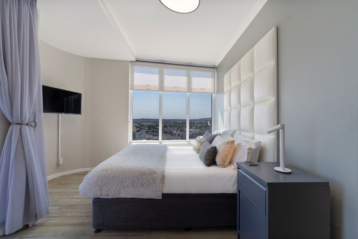 A bedroom with a large window, a bed, a bedside table, and a TV mounted on the wall.