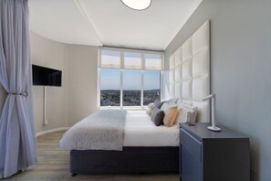 A bedroom with a large window, a bed, a bedside table, and a TV mounted on the wall. - Horizon Bay 903 Fresh Beachfront Hideaway (Bloubergstrand)