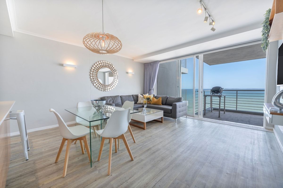 A modern living room with a glass dining table, white chairs, a grey sofa, and a large window overlooking the sea.