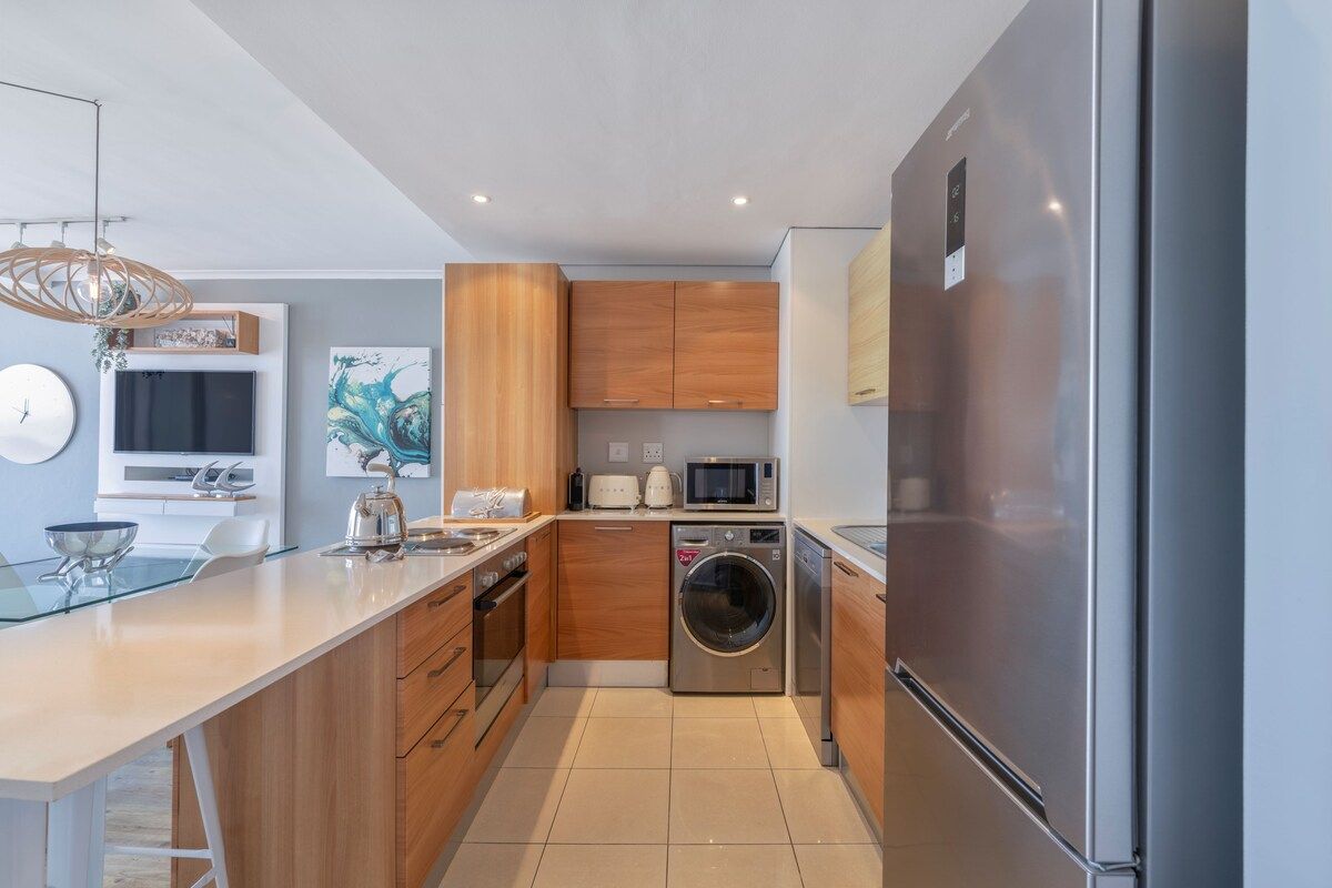 A modern kitchen with a stainless steel refrigerator, a built-in washing machine, and wooden cabinets.