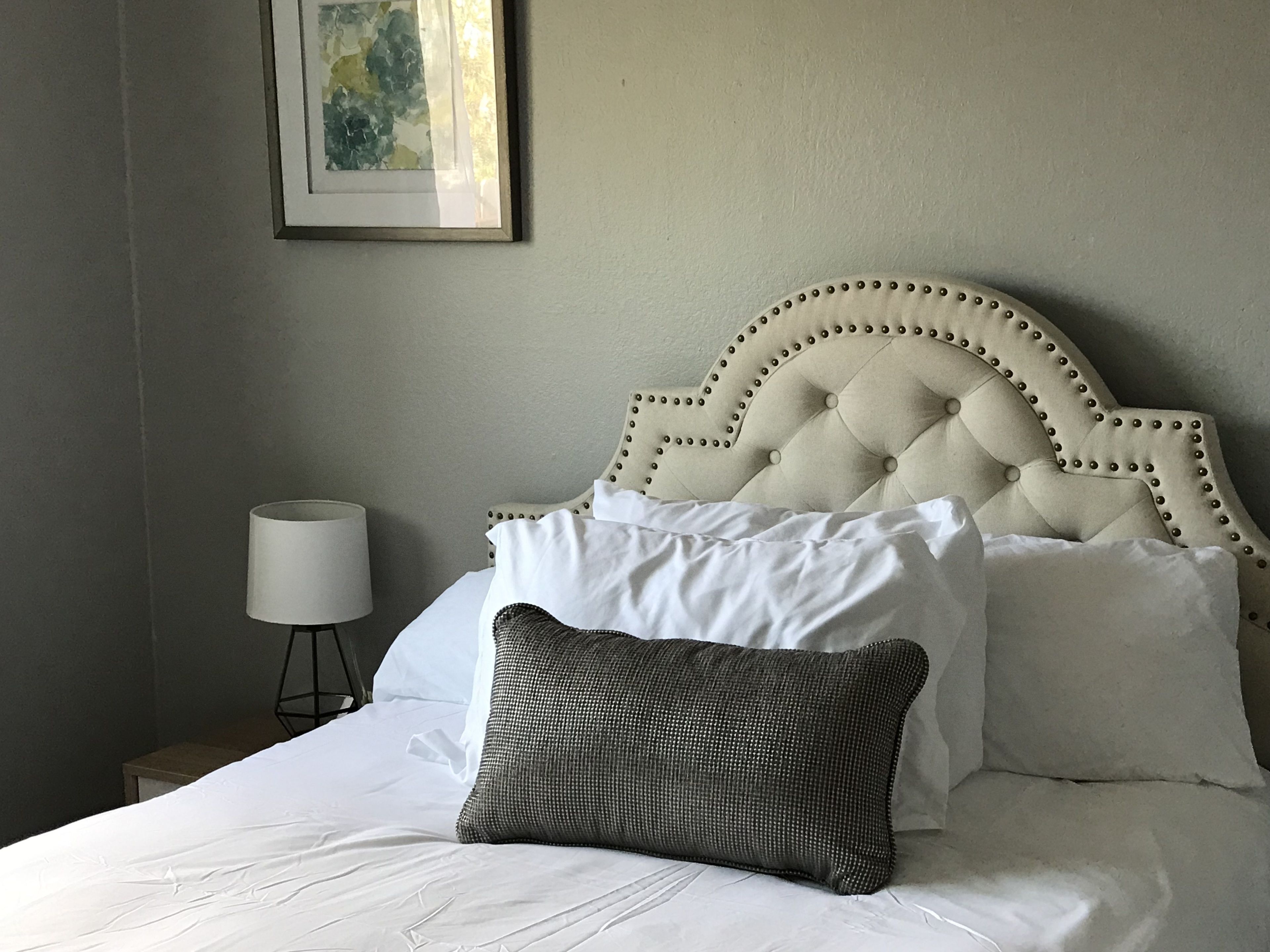 Uma cama bem arrumada com roupa de cama branca e cabeceira estofada, uma luminária de cabeceira e um quadro na parede.