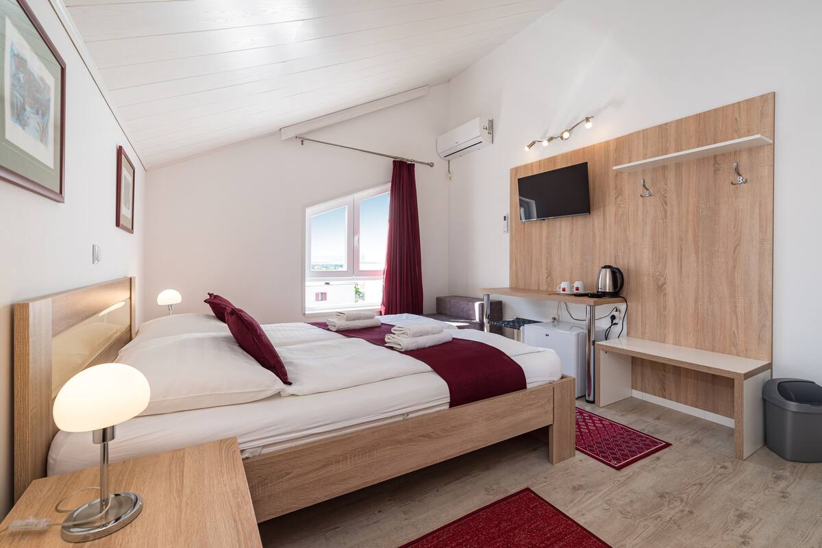 A modern bedroom with a wooden bed, a bedside table with a lamp, a TV mounted on the wall, and a small desk area.