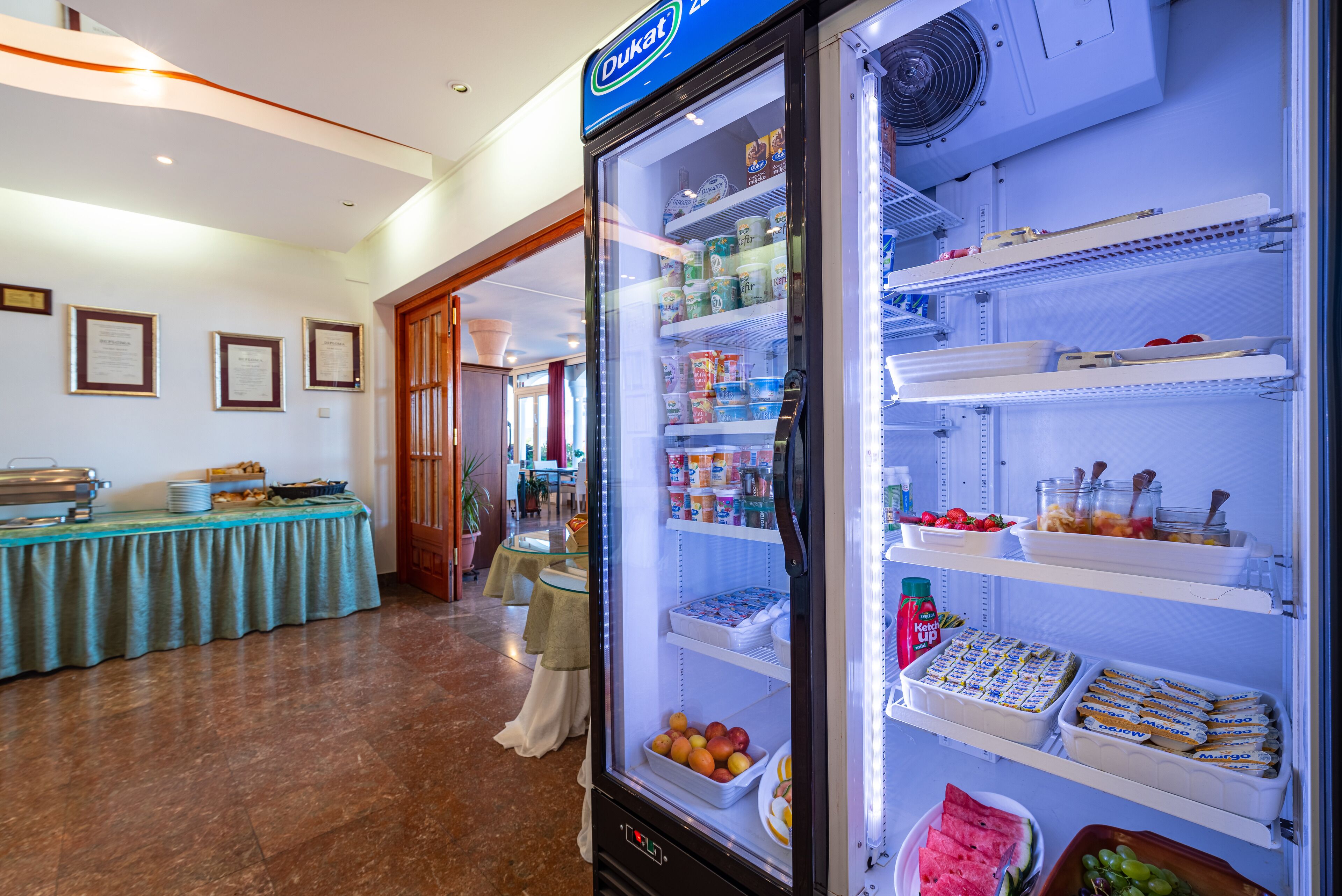 Mostrador refrigerado con diversos alimentos, una mesa de buffet con platos, y un comedor con mesas y sillas.