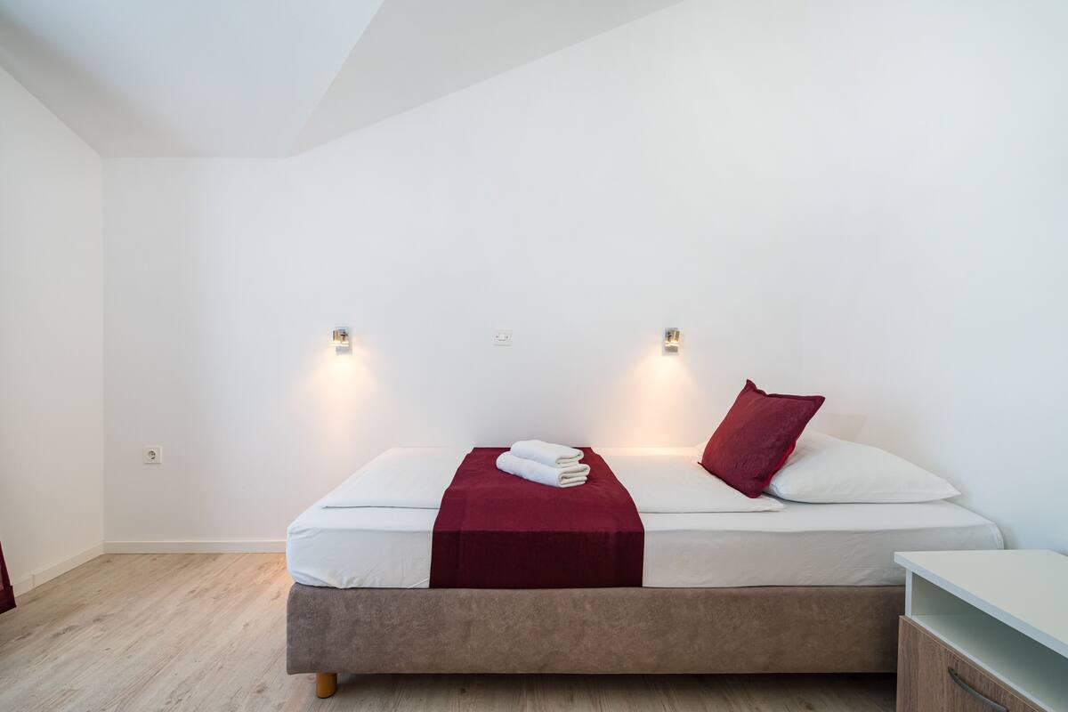 A neatly made bed with a red blanket and pillows, two wall-mounted lights, and a wooden floor.