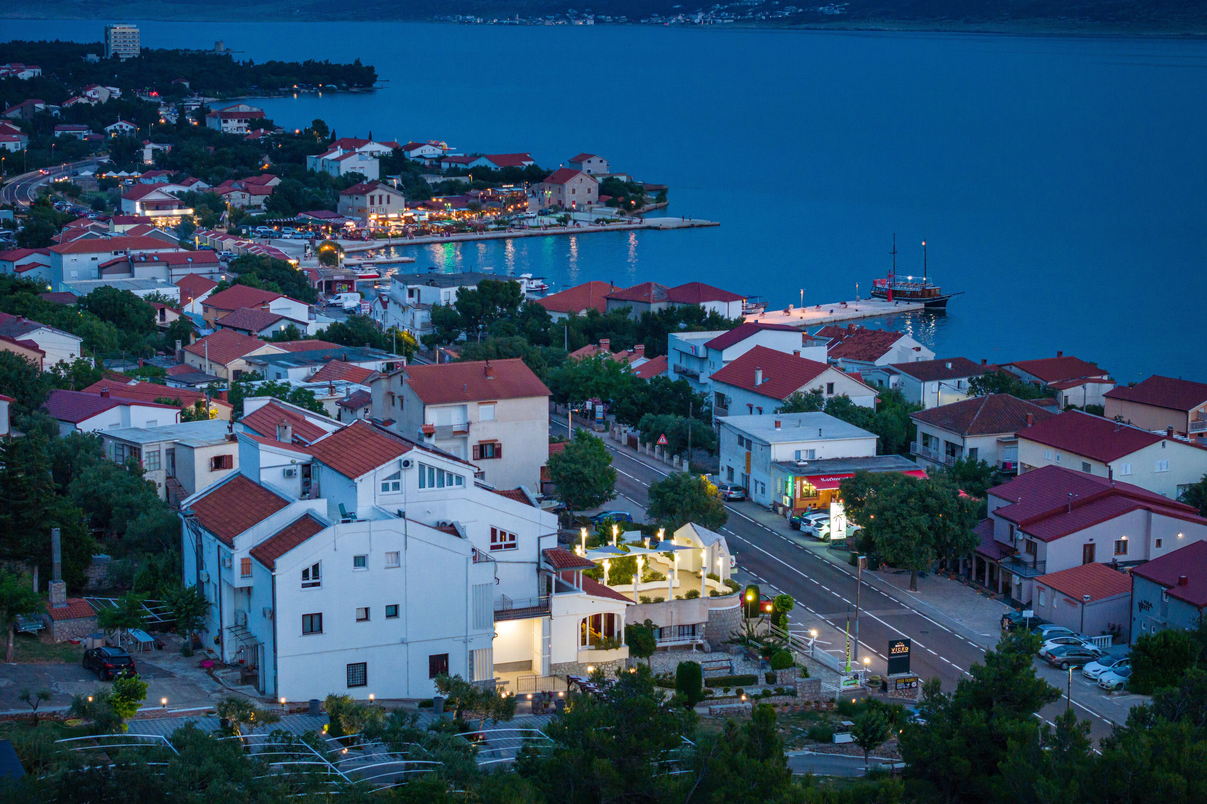 A coastal town with white buildings and red roofs, a marina, and a road leading to a brightly lit area.