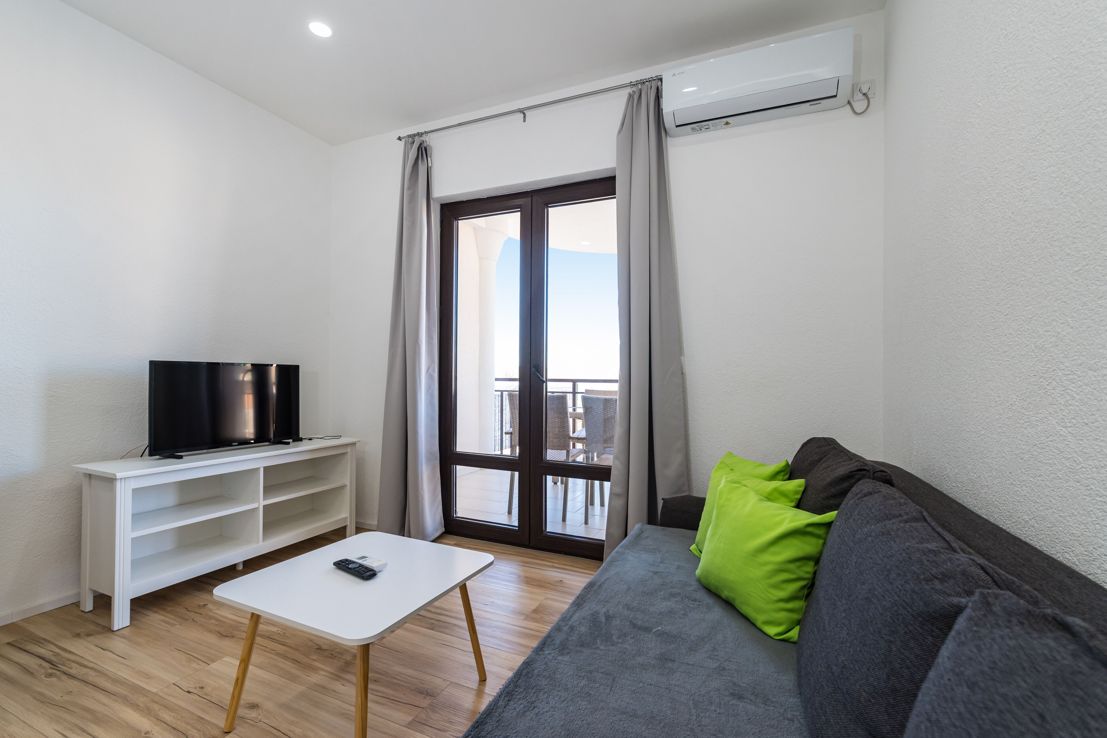 A modern living room with a flat-screen TV on a white stand, a grey sofa with a green pillow, a small white table, and a sliding glass door leading to an outdoor area.