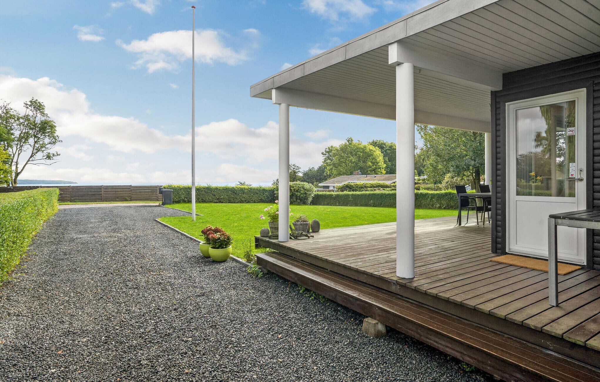 Un espace extérieur abrité, avec un chemin de gravier menant à une terrasse en bois, un coin salon avec une table et des chaises, et une vue sur un paysage verdoyant et des collines lointaines.
