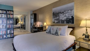 Una habitación de hotel con una cama grande, una estantería, un escritorio con lámpara y un cuadro de un paisaje urbano.