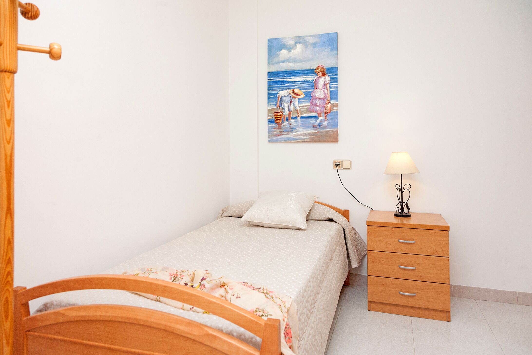 Une chambre avec un lit en bois, une table de chevet avec une lampe, un tableau représentant une scène de plage et un mur blanc.