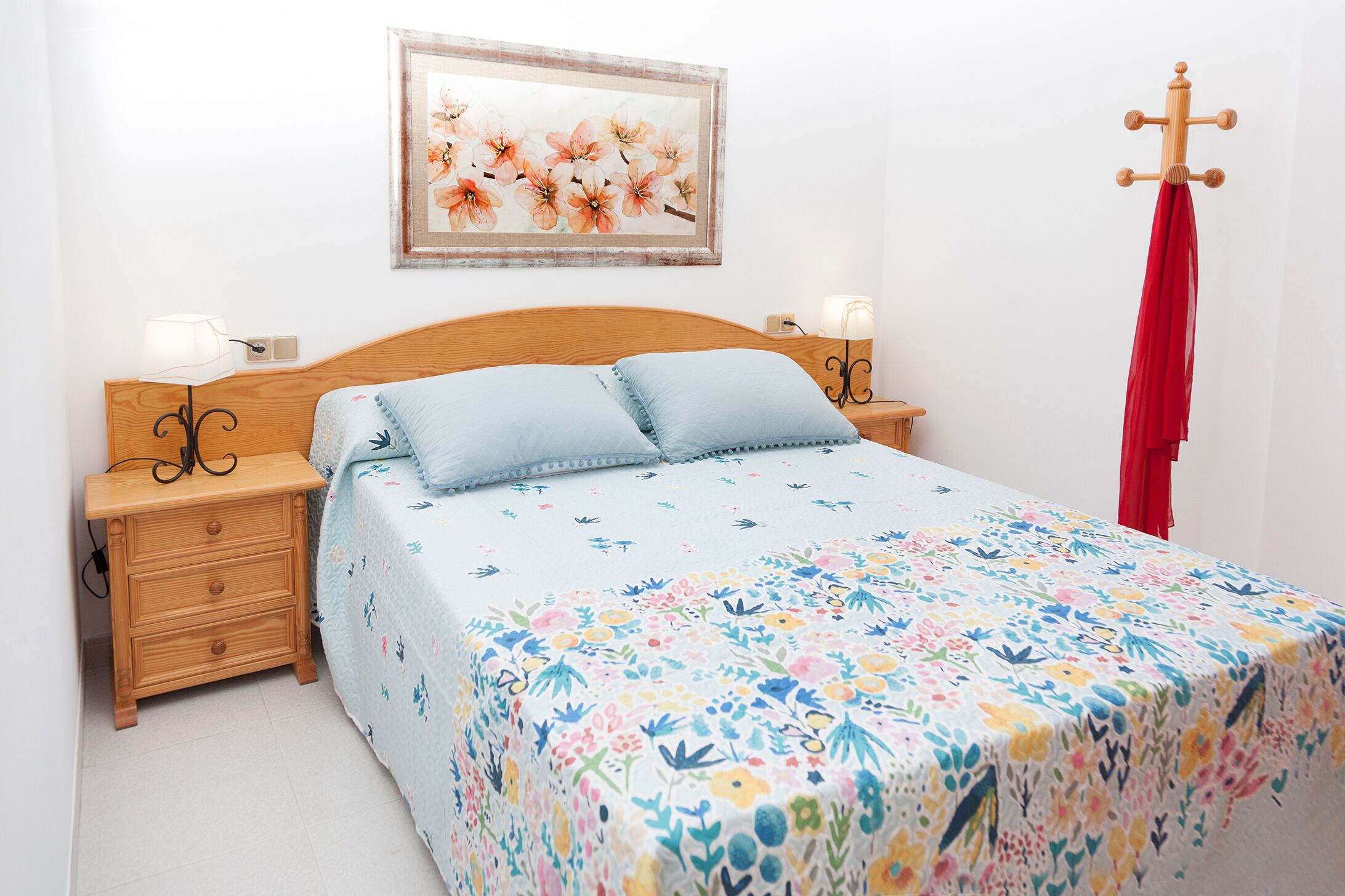 A bedroom with a floral bedspread, a wooden headboard, two bedside tables with lamps, a red towel hanging on a wooden rack, and a framed floral painting on the wall.