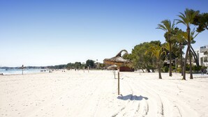 Ein weiter Sandstrand mit wenigen Palmen und einem Gebäude mit einem Reetdach.