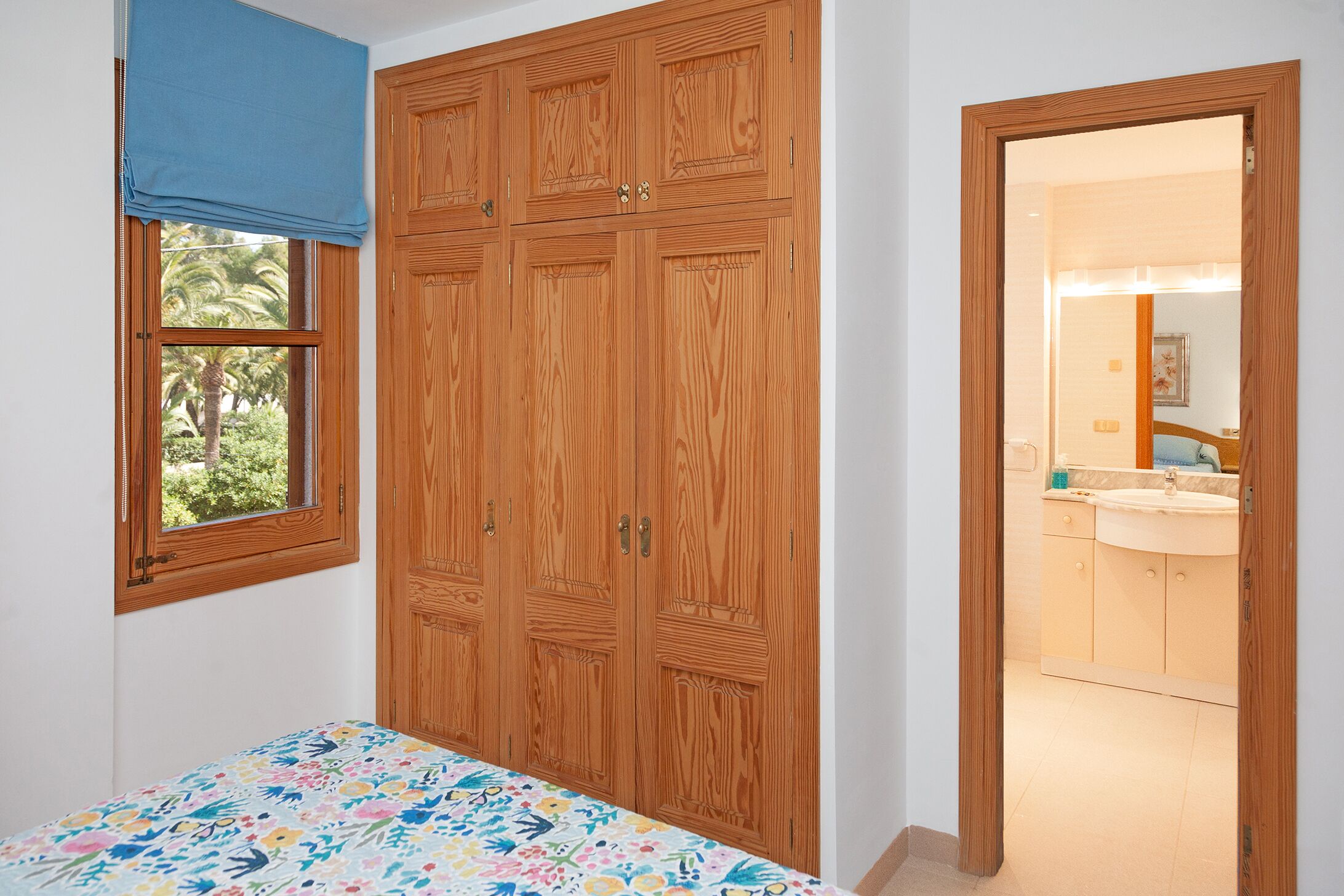 A bedroom with a wooden wardrobe, a window with a blue curtain, and a view of a bathroom with a sink and mirror.
