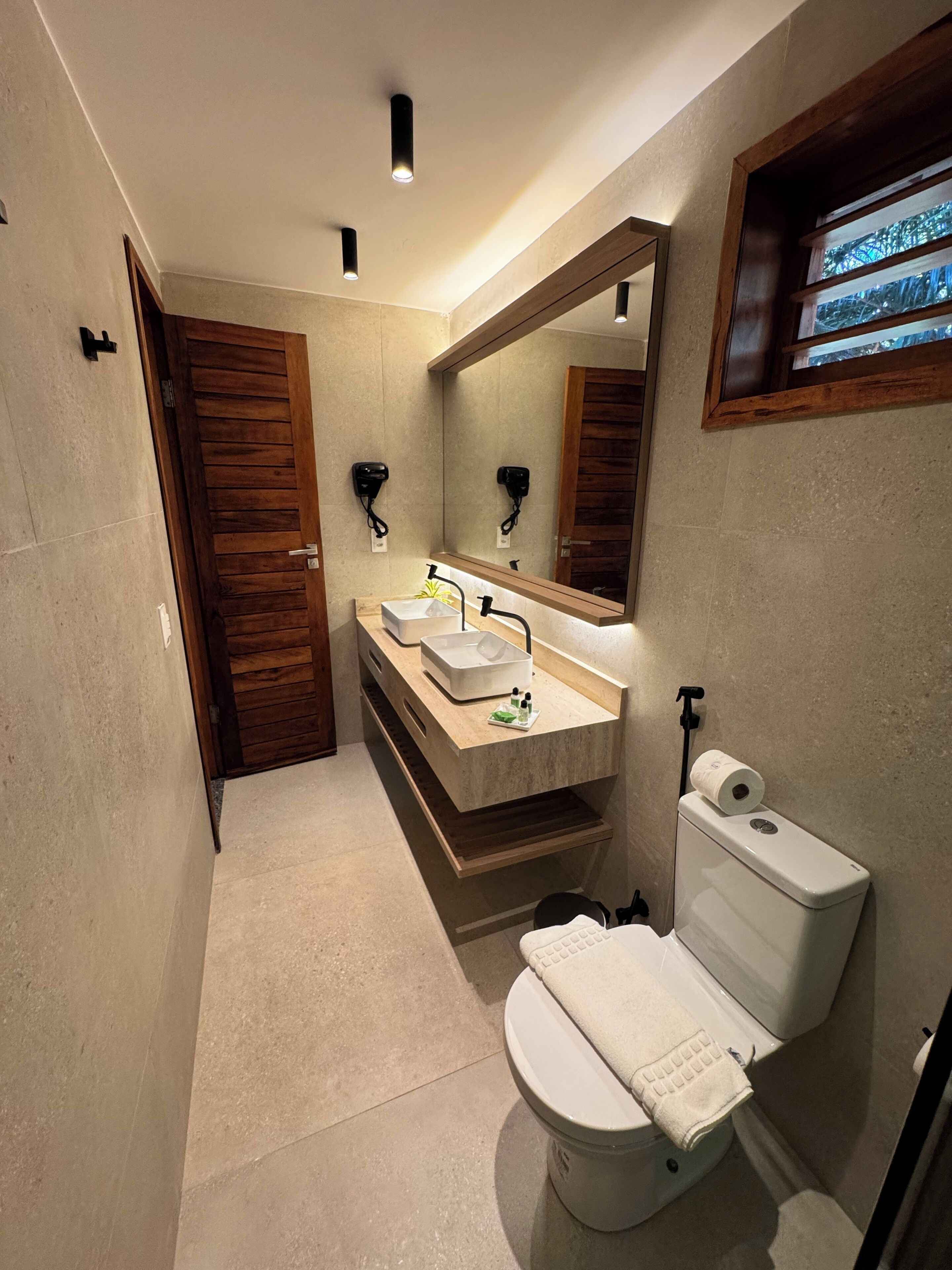 A modern bathroom with a wooden door, a large mirror, a sink with a towel, and a toilet.