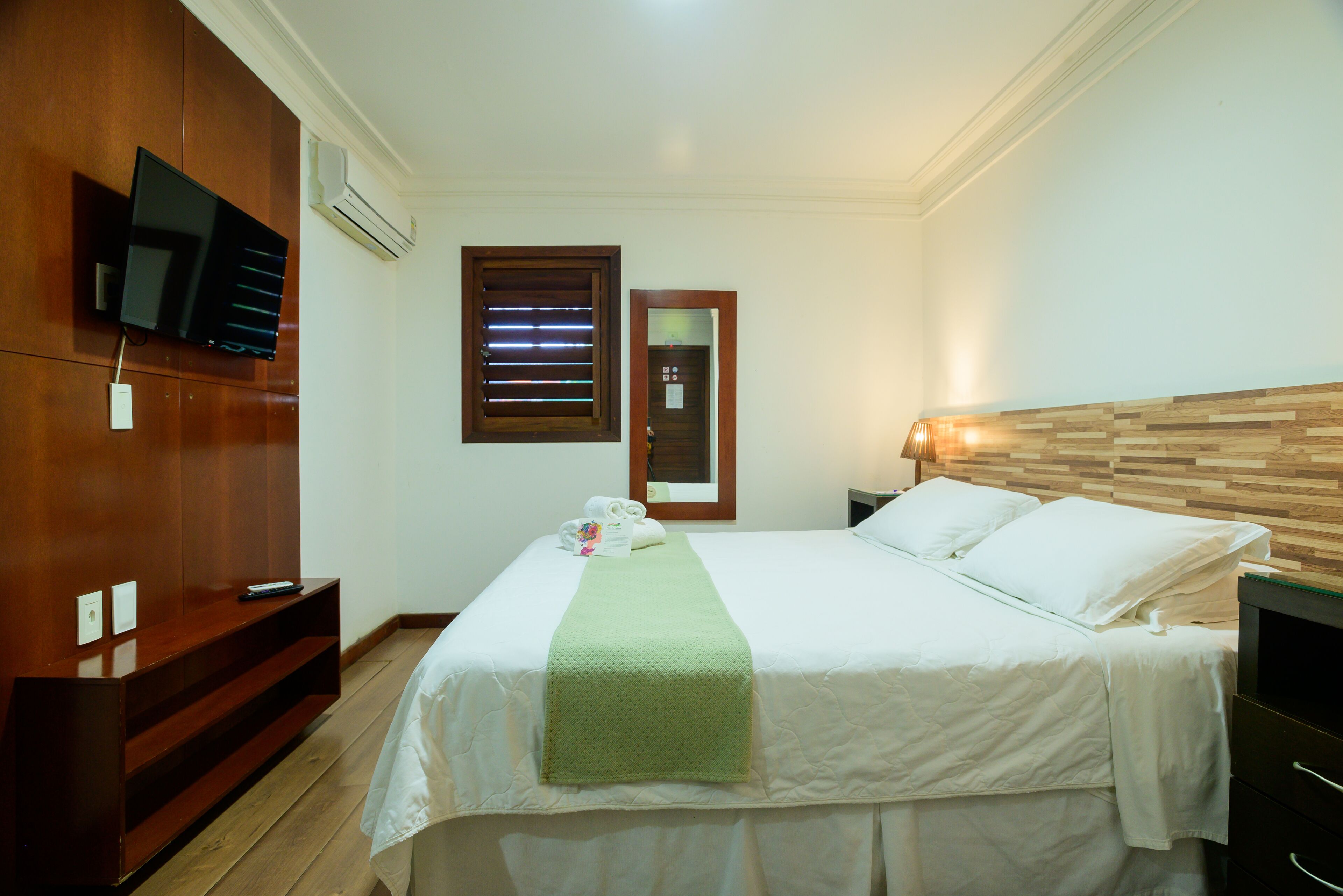 Hotelkamer met een bed, een aan de muur bevestigde televisie, een houten hoofdeinde en een nachtkastje met een lamp.