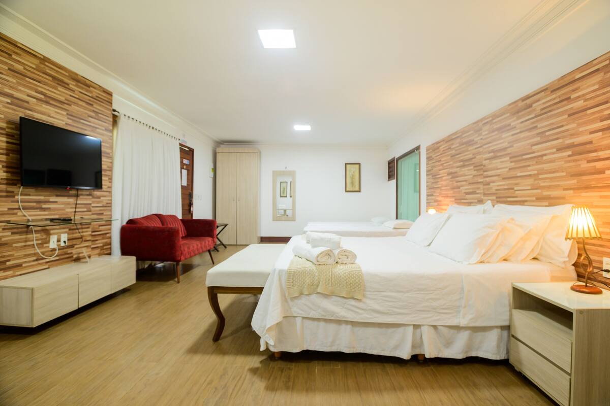 A hotel room with a wooden wall, a flat-screen TV, a red chair, two beds, a bedside table with a lamp, and a bench.