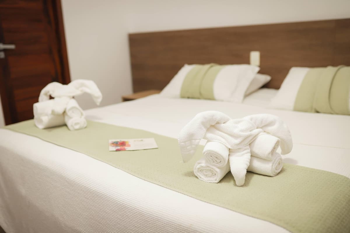 A neatly made bed with rolled towels and a green towel bar, a wooden headboard, and a white wall.