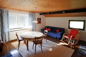Una sala de estar acogedora con una mesa redonda, sillas de madera, un sofá azul con cojines rojos, un sillón mecedora y una ventana con cortinas estampadas.