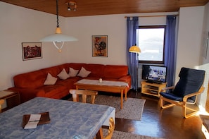 Una sala de estar con un sofá rojo, una mesa de centro de madera, una mesita azul estampada, una silla de madera, un televisor y una ventana con cortinas azules.
