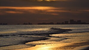 Un coucher de soleil sur une étendue d’eau, avec des vagues et le profil d’une ville lointaine à l’horizon.
