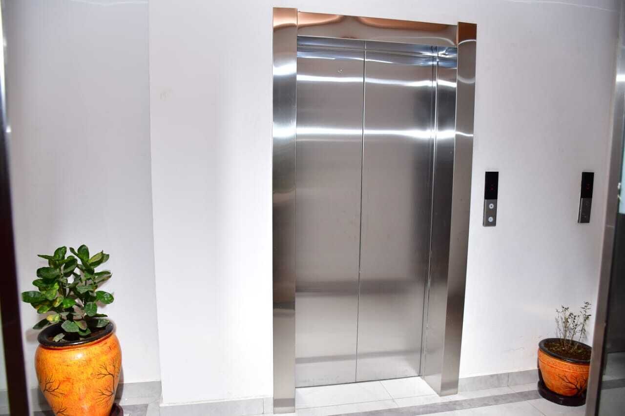 Elevador moderno com acabamento metálico, ladeado por duas plantas em vasos.