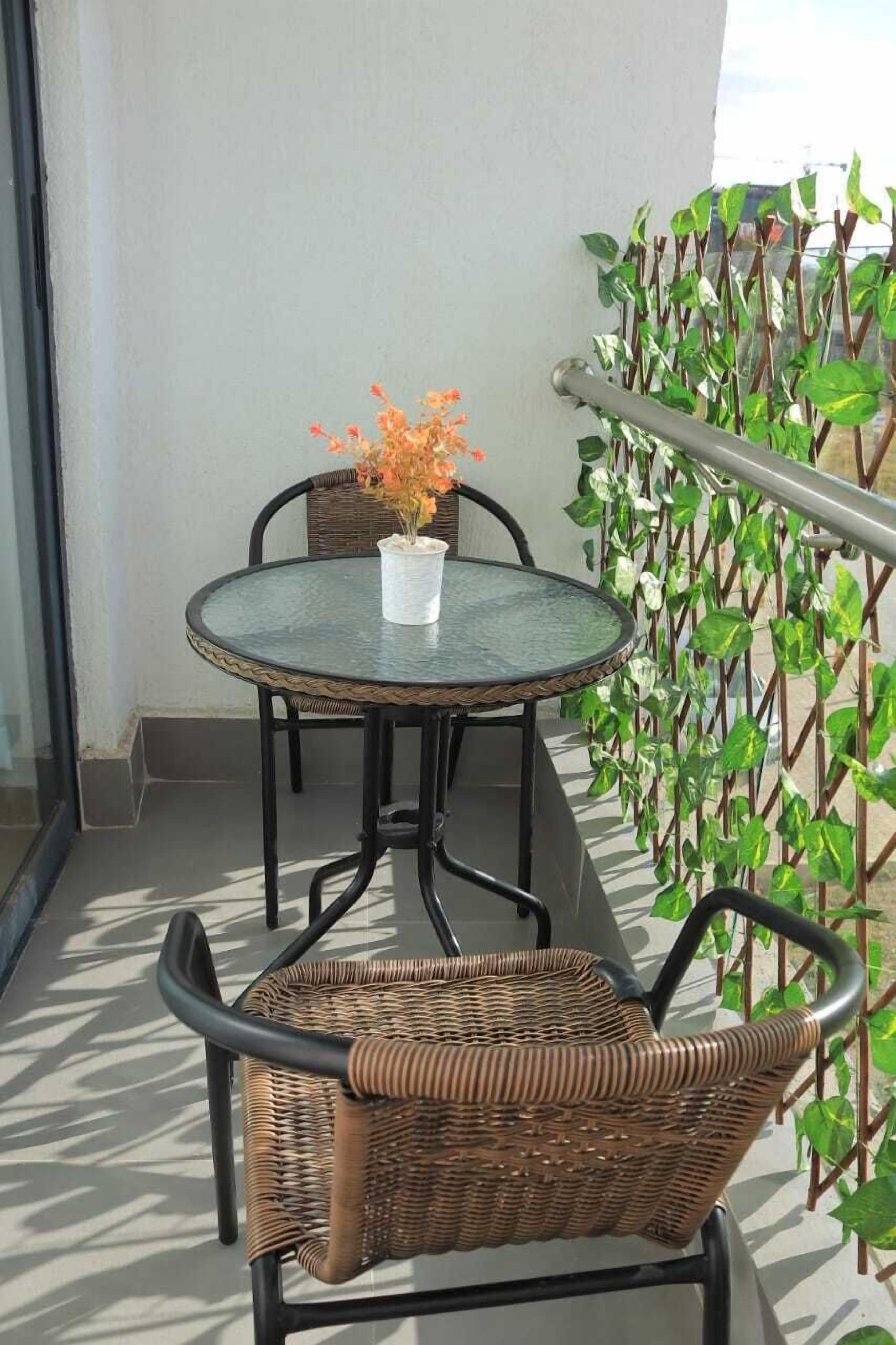 A balcony with a round table and two wicker chairs, a potted plant, and a metal railing.