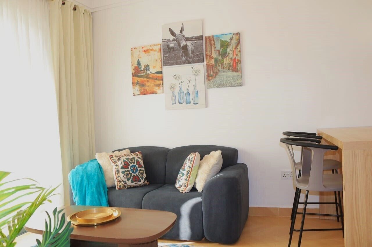 A living room with a grey sofa, a small round table, a bar stool, and three wall-mounted artworks depicting a horse, flowers, and a street scene.