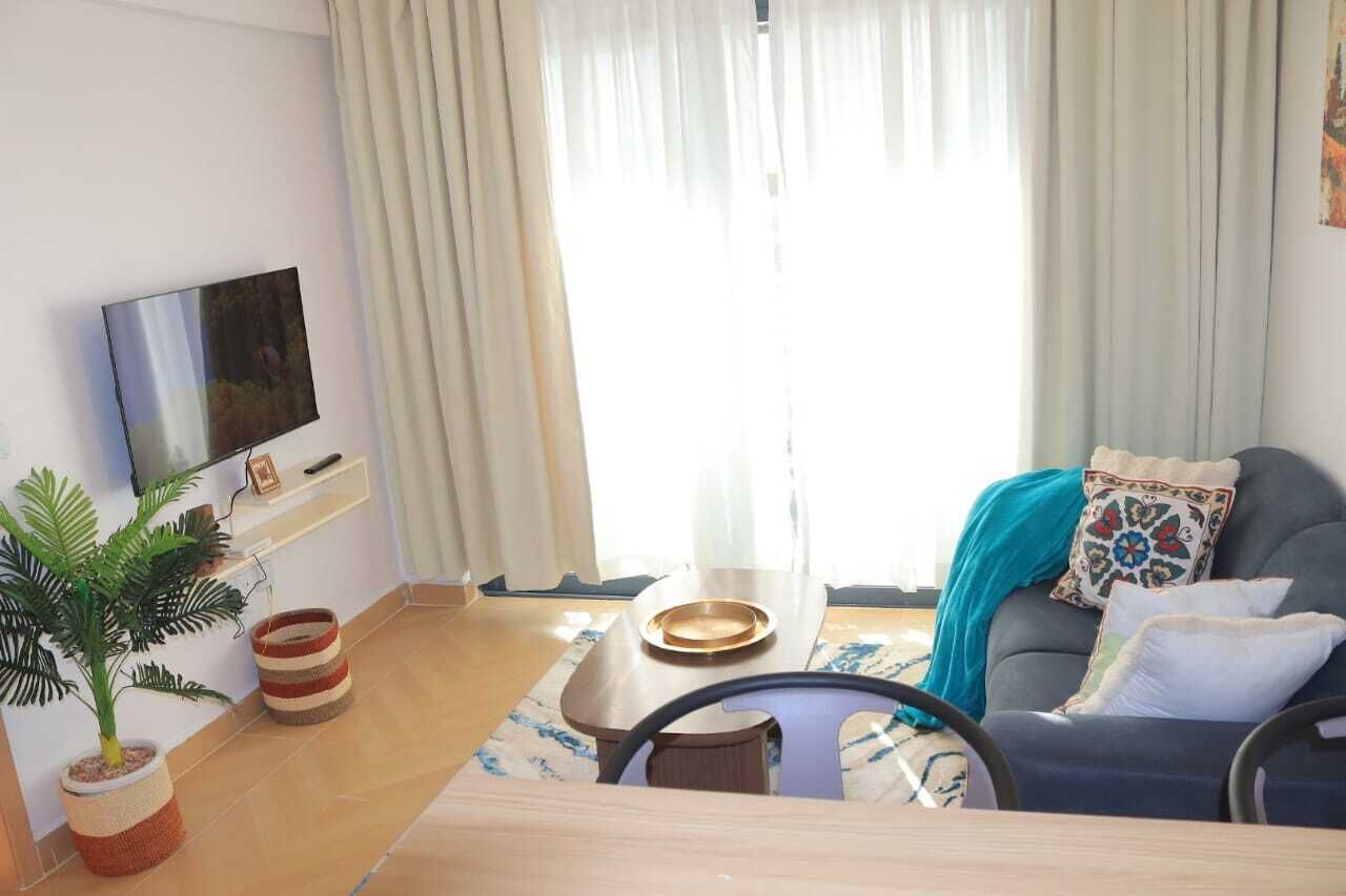 A living room with a sofa, a coffee table, a TV mounted on the wall, a potted plant, and a window with curtains.