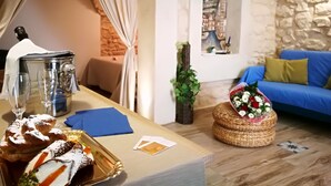 Ein gemütliches Zimmer mit Esstisch, gedeckten Speisen, Gebäck und einer Kaffeekanne, einem blauen Sofa und einem dekorativen Korb mit Blumen.