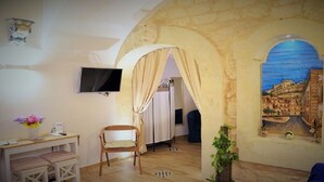 Ein Zimmer mit einer Steinswand, einem Bild von einer Küstenstadt, einem Schreibtisch mit Stuhl und einem an der Wand montierten Fernseher.