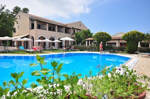 A swimming pool with lounge chairs and umbrellas, surrounded by a building with balconies and a tiled roof. - Acharavi Garden (Corfu)
