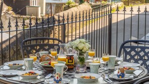 Una mesa de desayuno preparada con platos, tazones y vasos de jugo de naranja, con vistas a una zona costera pintoresca con una valla y la vista de barcos en el agua.