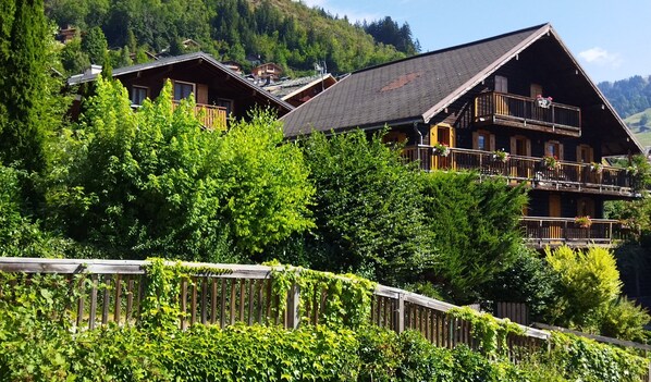 En byggnad i chaletstil med flera balkonger, omgiven av frodig grönska och med en imponerande bergskedja i bakgrunden.