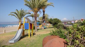 Un parque infantil con un tobogán y estructuras para escalar, palmeras y una playa al fondo.