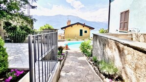 Un chemin de pierres mène à une maison jaune qui offre une vue sur la montagne, et qui est entourée de verdure et d’une piscine.