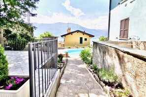 A stone pathway leading to a yellow house with a mountain view, surrounded by greenery and a swimming pool.