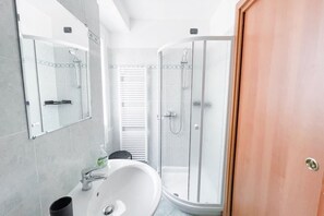 Ein Badezimmer mit weißem Waschbecken, einem Spiegel, einer Duschkabine aus Glas und einer Holztür.