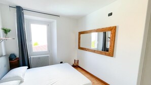 Ein Schlafzimmer mit Bett, einem Vorhangfenster, Heizkörper, Holzboden, einem kleinen Stuhl, einem Nachttisch mit Lampe und einem Spiegel an der Wand.