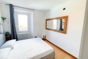 A bedroom with a bed, a window with curtains, a radiator, a wooden floor, a small chair, a nightstand with a lamp, and a mirror on the wall.