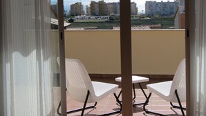 Ein Balkon mit zwei weißen Stühlen und einem kleinen Tisch, der auf ein Wohngebiet mit Gebäuden und einem klaren Himmel blickt.