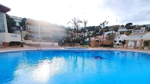 Ein Pool mit gewölbter Kante, umgeben von einem hellen Deck. Im Hintergrund sind Gebäude mit Balkonen und etwas Grünwerk zu sehen.
