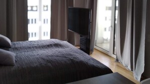 Habitación de hotel con cama, televisor y mesa. Hay cortinas en las ventanas y una vista de edificios desde el exterior.