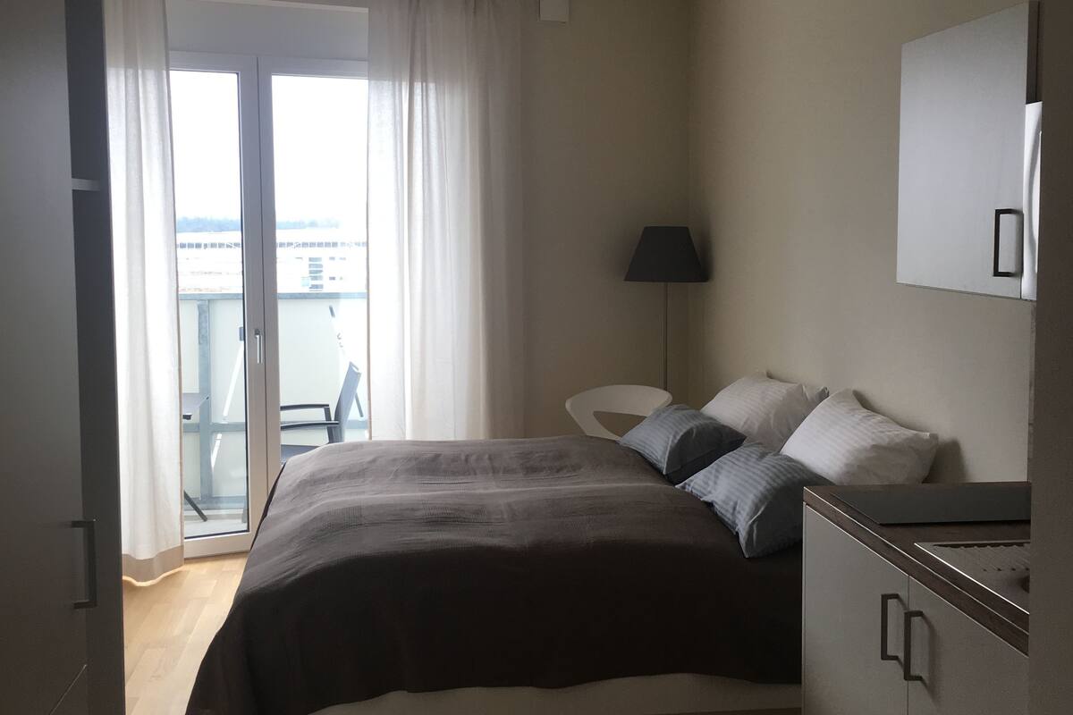 A bedroom with a bed, a nightstand, a lamp, and a view of the outside.