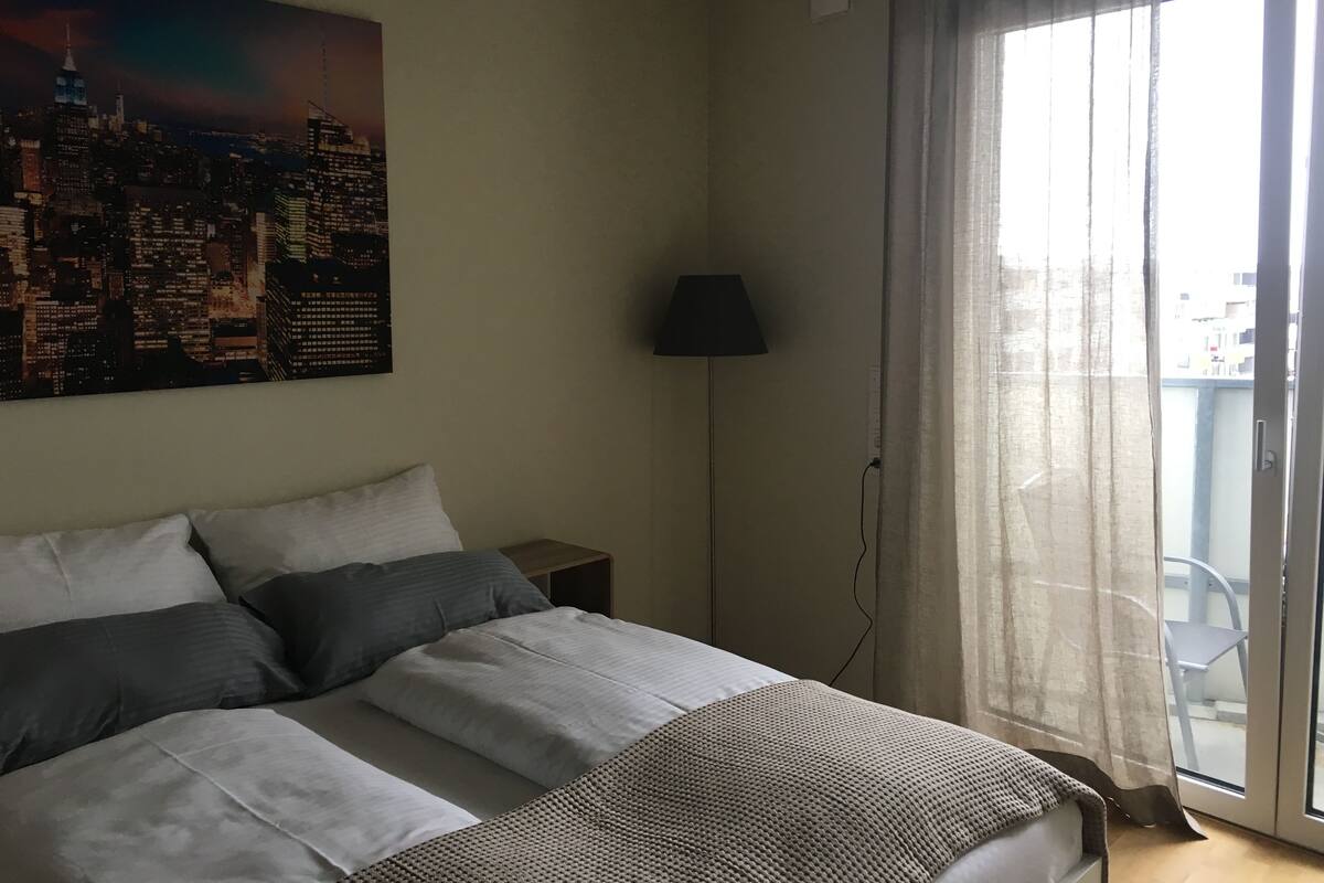 A bedroom with a bed, pillows, a lamp, and a cityscape painting on the wall.