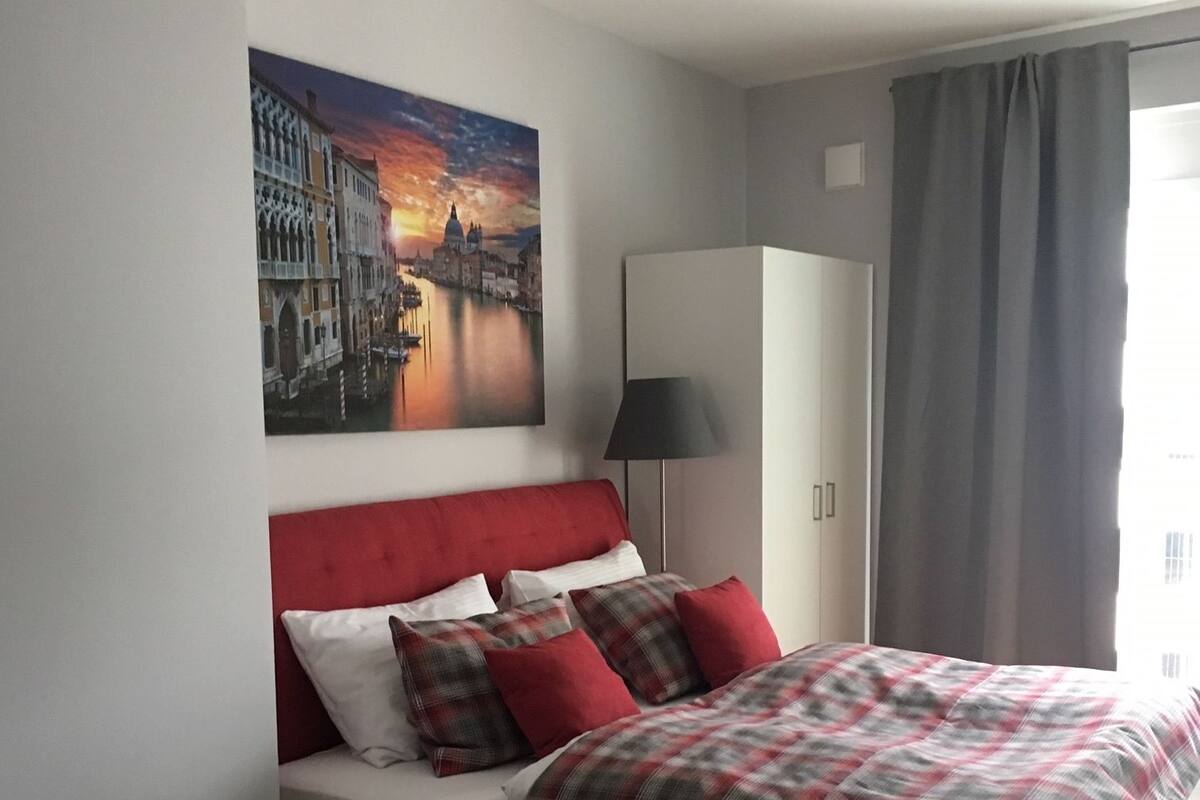 A hotel room with a bed, a desk, a chair, a lamp, and a painting of a cityscape.