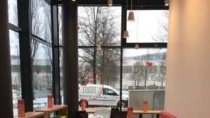 Un interior de restaurante con ventanas altas, mesas de madera, sillas coloridas y una zona de estar con cojines rojos.
