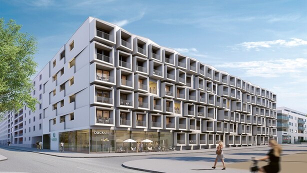 Un edificio moderno con una fachada de balcones enrejados, un escaparate con letrero y un espacio peatonal con mesas al aire libre.