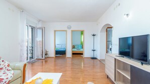 Ein großzügiges Wohnzimmer mit einer Couch, einem Couchtisch mit Dekoration, einem Fernseher und einem Kamin. Zwei Türen führen in andere Räume.
