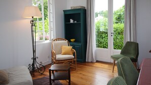 Una habitación con piso de madera, un sillón verde, una silla de mimbre, una mesita, una lámpara de pie y un mueble verde.